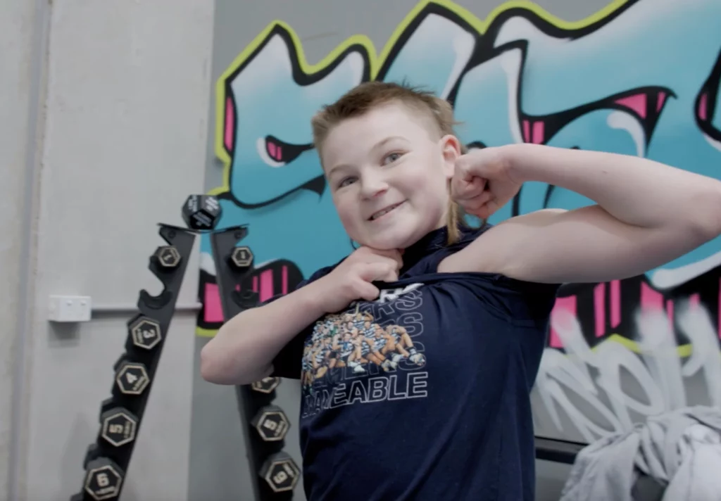 Smiling teenage boy flexes arm muscles while working individual therapy with peer mentor Will