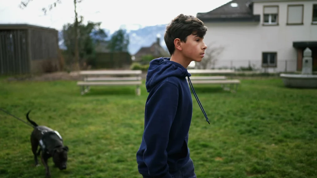 Teenage boy in blue hoodie looking thoughtful outdoors with a dog nearby" Teenage boy in blue hoodie looking thoughtful outdoors with a dog nearby